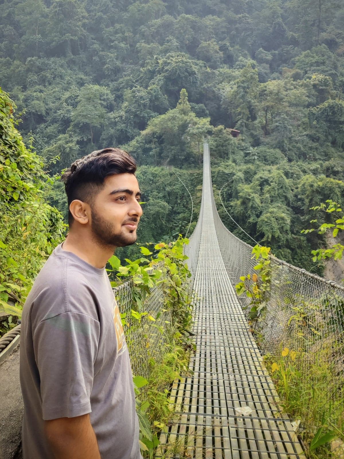 Sanchit Pandey standing at the entrance of a long suspension bridge over a forested valley