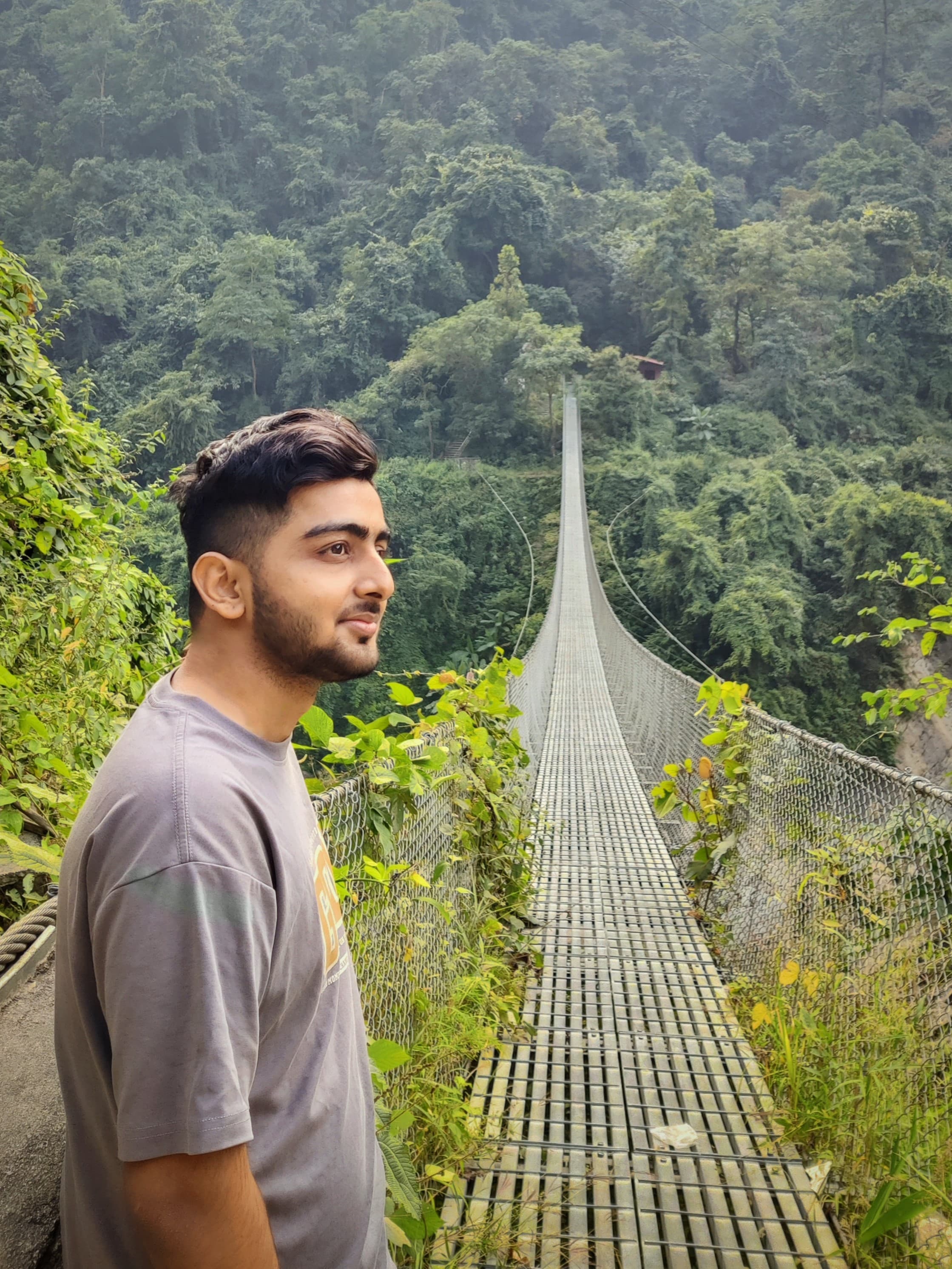 Sanchit Pandey standing at the entrance of a long suspension bridge over a forested valley